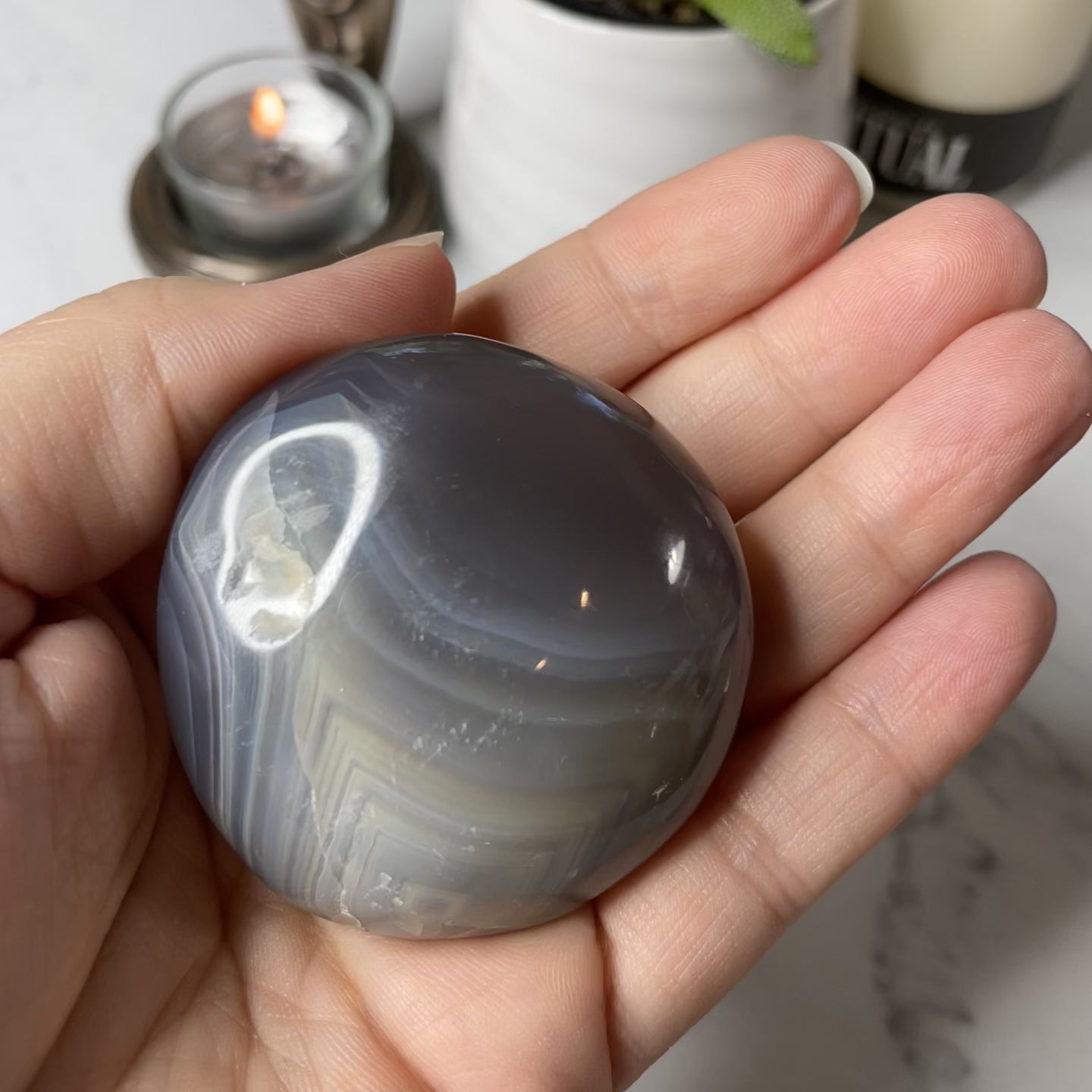 Freya's Haven | Metaphysical & Crystal shop | Three blue and white banded palm stones of Orca Agate are stacked in a white Selenite bowl on a white marble tile. There are candles and a pot in the background.