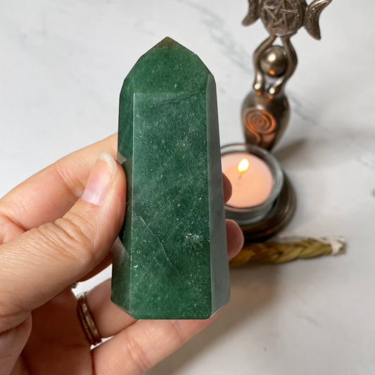 A Green Aventurine tower is held in a woman's hand over a white marble base with a bronze female Goddess tea light holder in the background