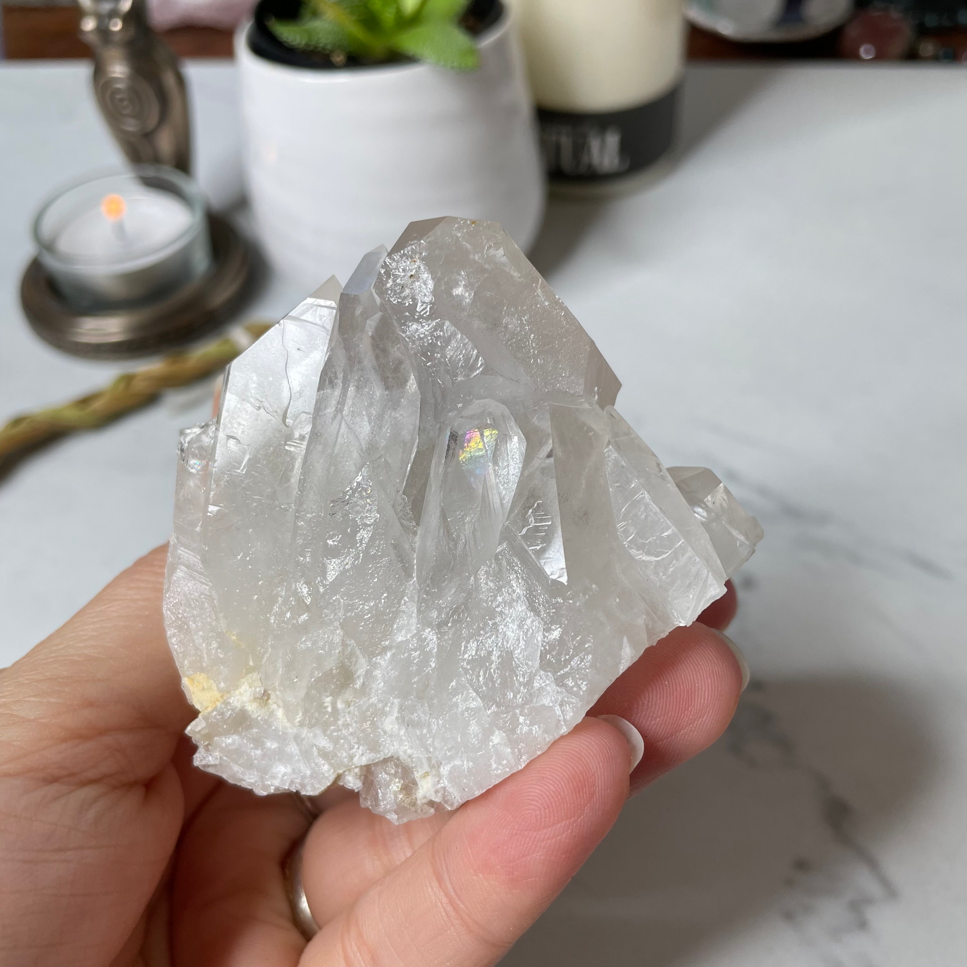 Freya's Haven | Metaphysical & Crystal shop | A close up photo of a Clear Quartz Cluster held in a woman's hand with candles in the background. A rainbow can be seen inside one of the crystal's points.