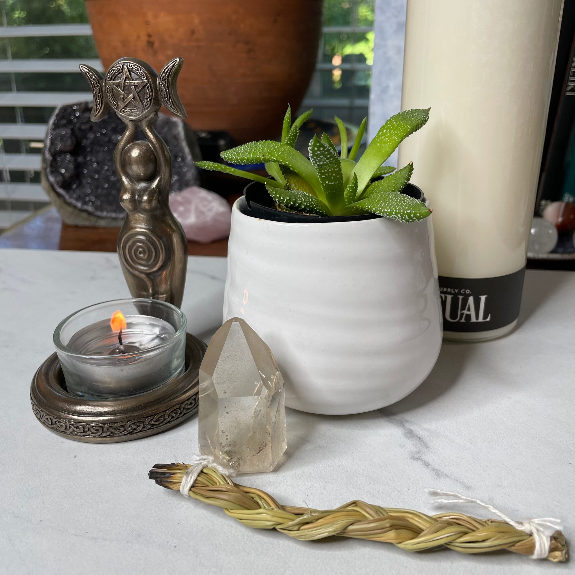 Freya's Haven | Metaphysical & Crystal shop | A pale yellow stone called Citrine has been polished into a standing point. It is displayed with a succulent in a white pot, a goddess tea light candle holder with lit candle and a braid of sweet grass on a white marble tile. there are other crystals, a plant and books in the background.