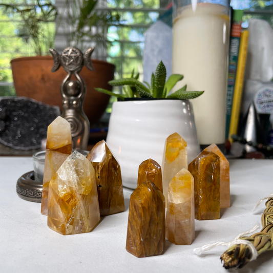 Freya's Haven | Metaphysical & Crystal shop | A collection of nine golden stones called Golden Healer have been polished into standing points. They are displayed with a succulent in a white pot, a goddess tea light candle holder with lit candle and a braid of sweet grass on a white marble tile. there are other crystals, a plant and books in the background.