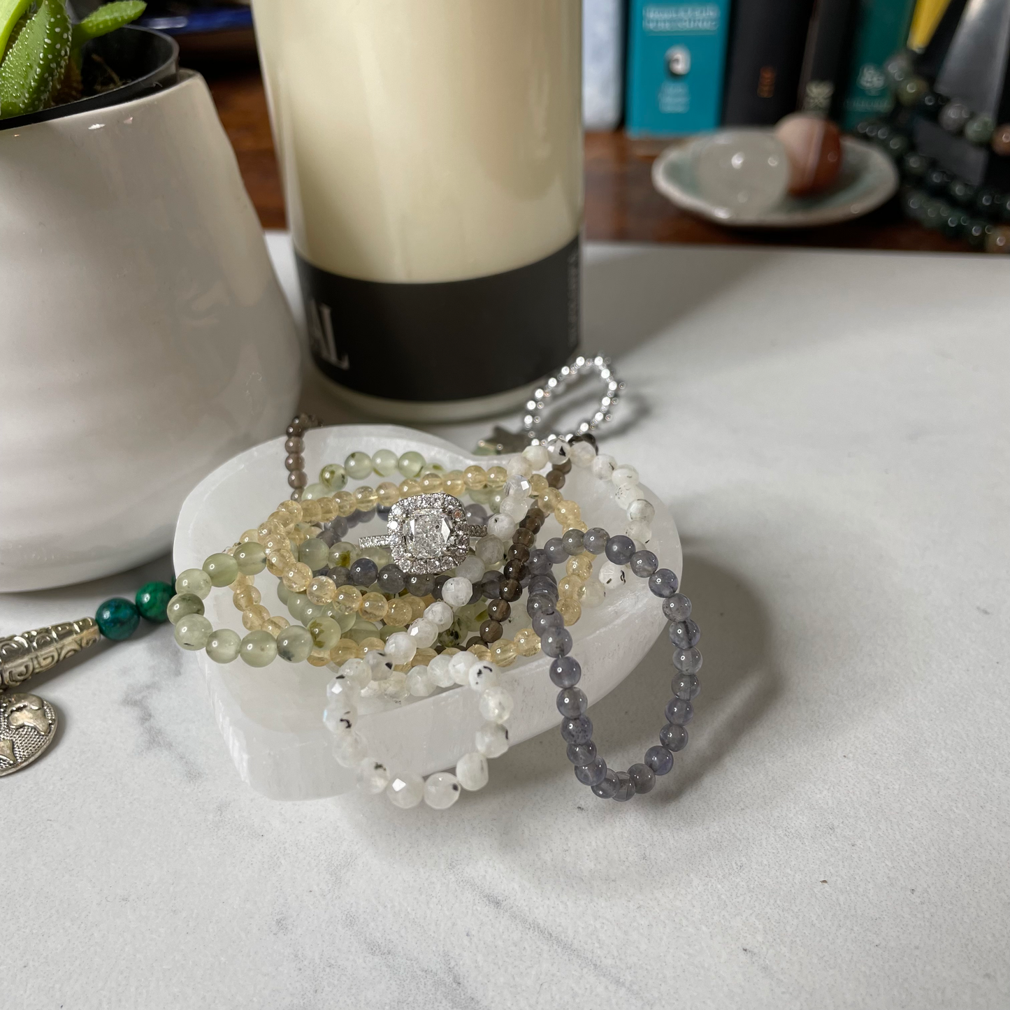Freya's Haven | Metaphysical & Crystal shop | A  heart-shaped Selenite bowl is displayed with a succulent in a white pot, a goddess tea light candle holder with lit candle with a few crystal bracelets and diamond engagement ring displayed inside the bowl.